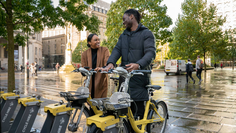 Two people smiling in Manchester City Centre as they are unlocking a cycle hire bike