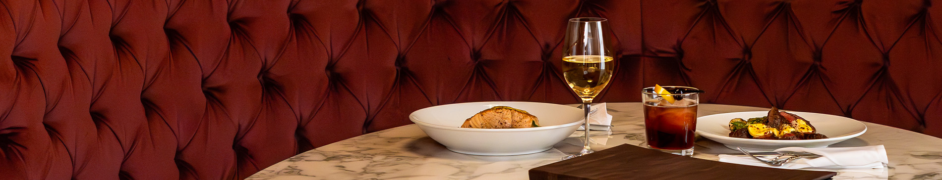 Two plates of food with drinks on a marble table with red fabric bench seat behind. 