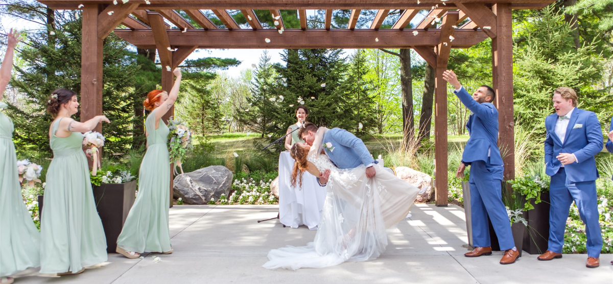 Groom dips and kisses bride as guests throw petals at an outdoor wedding ceremony.