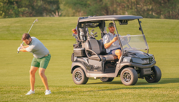Male golfer swinging while his friend waits in a golf cart. 