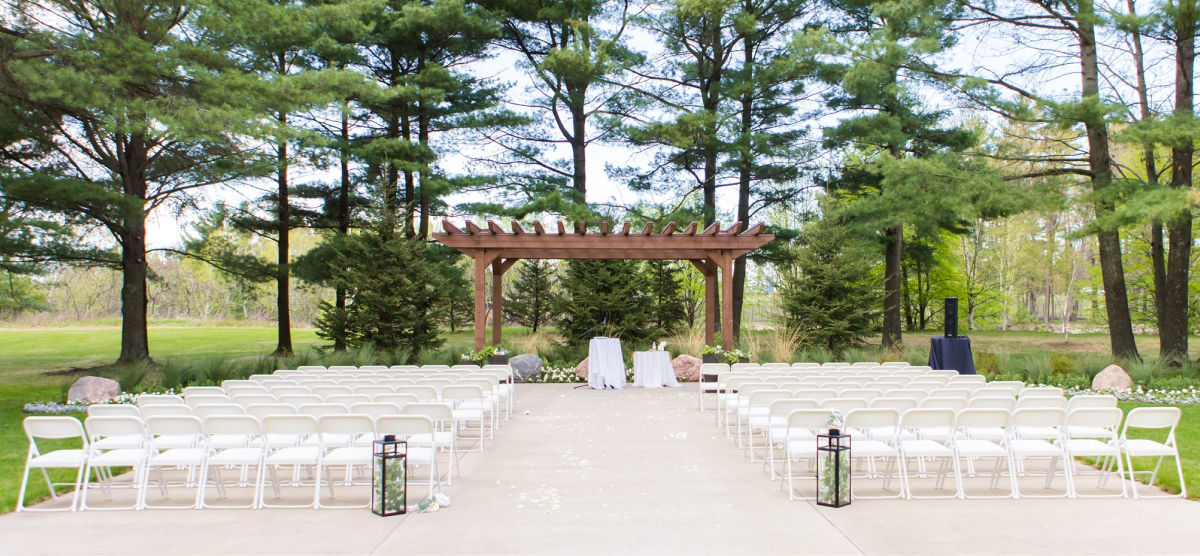 Outdoor wedding ceremony setup with rows of white chairs, a paved aisle, and a wooden pergola.
