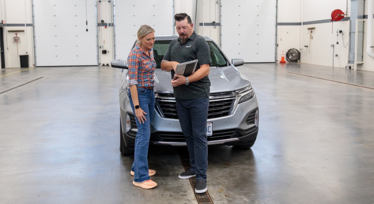 Two people looking at a laptop in front of a silver car in a garage.