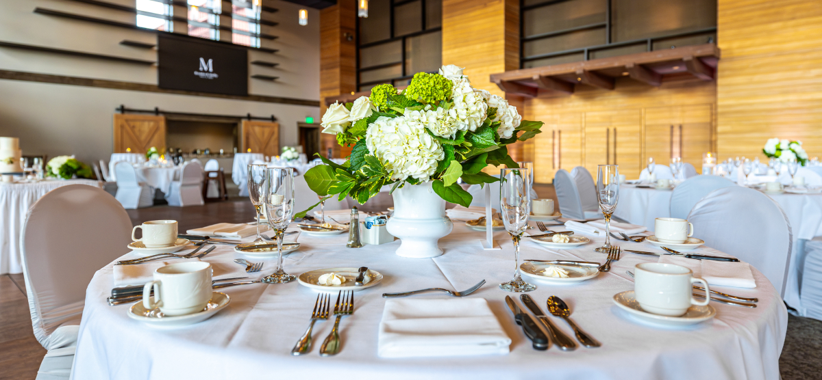 Event table set with white linens, a white and green floral centerpiece, and place settings.