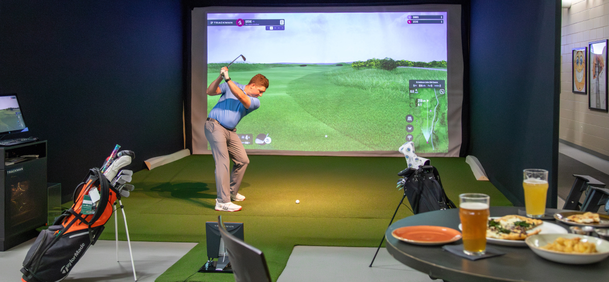 Man in blue shirt swinging a golf club in a golf simulator with a projected course.