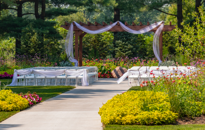 Outdoor wedding ceremony with white chairs, a paved aisle, and a fabric-draped wooden pergola.