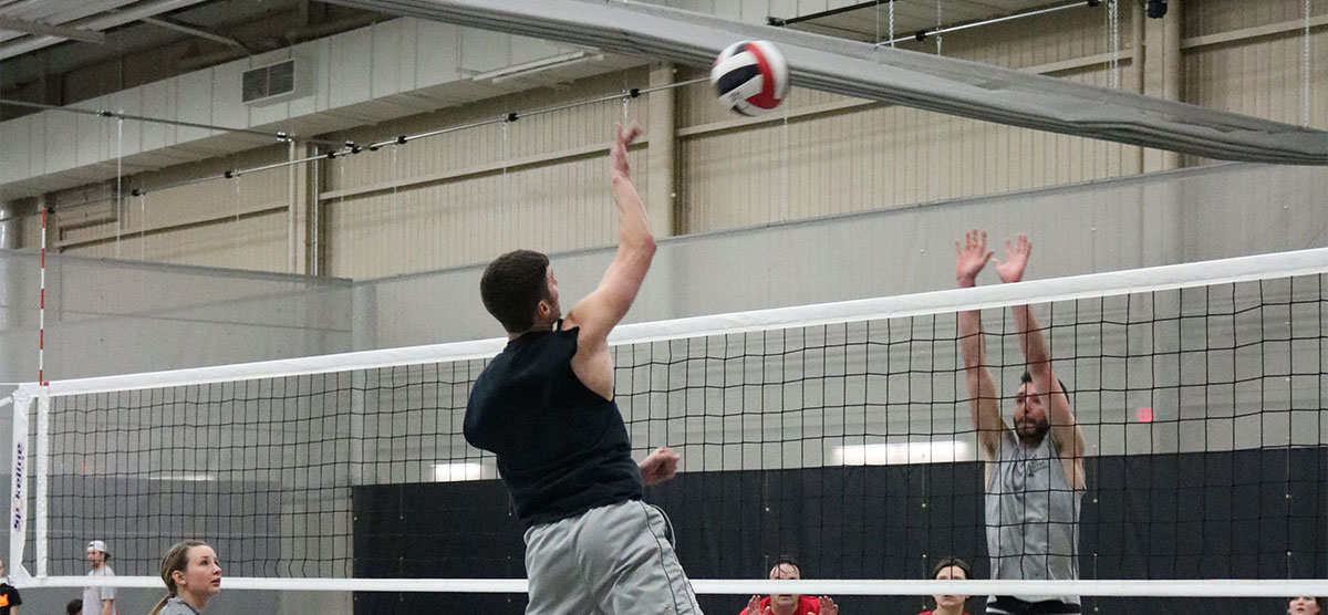 Player hitting a volleyball and another blocking at the net during a game in the fieldhouse