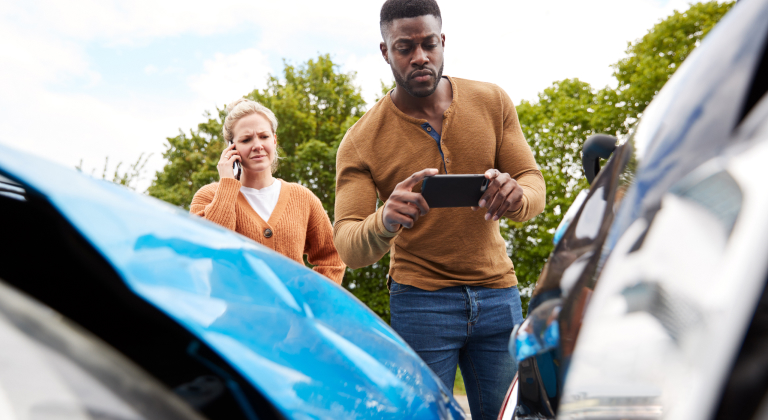 Man taking photo of car damage with phone, woman on phone looking concerned.