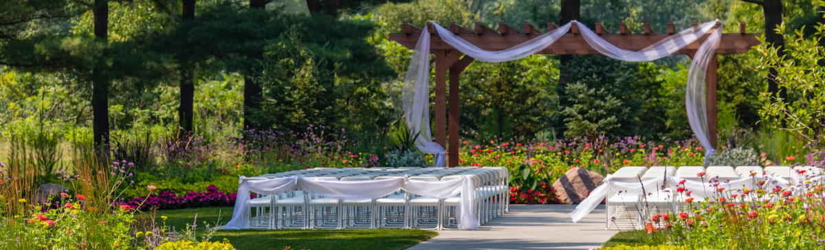 Outdoor wedding ceremony with white chairs, a paved aisle, and a fabric-draped wooden pergola.
