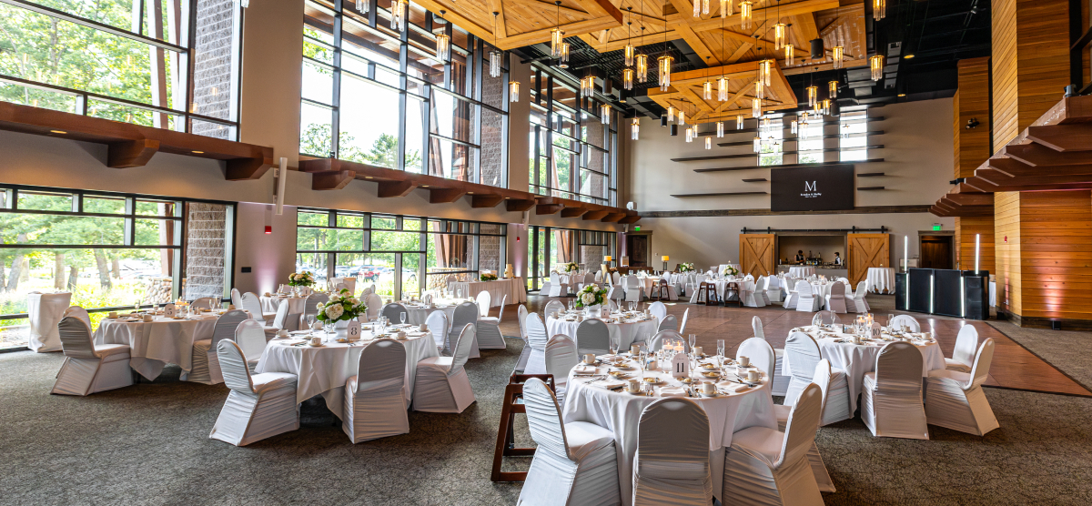 Large event room with many round tables set for a reception, featuring white linens and floral centerpieces.