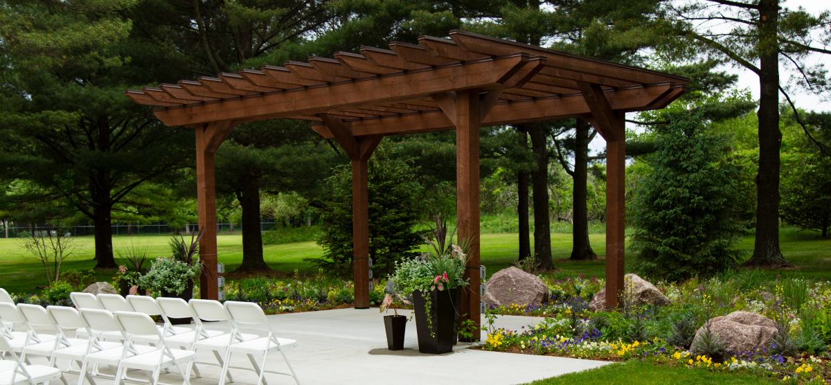 Outdoor event space with a wooden pergola, white chairs, and garden landscaping.