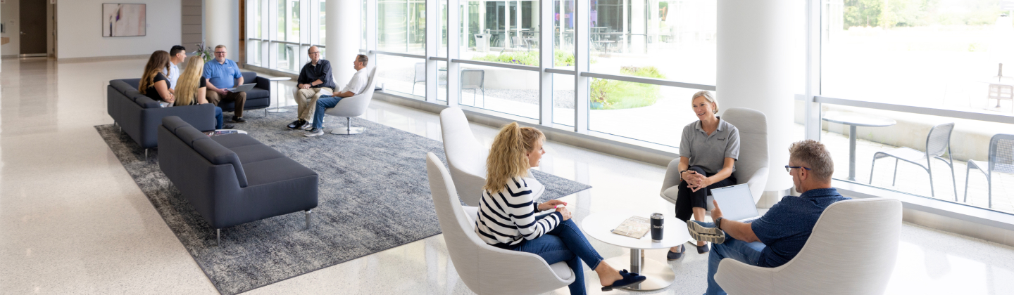 Employees seated and talking in front of a wall of windows. 