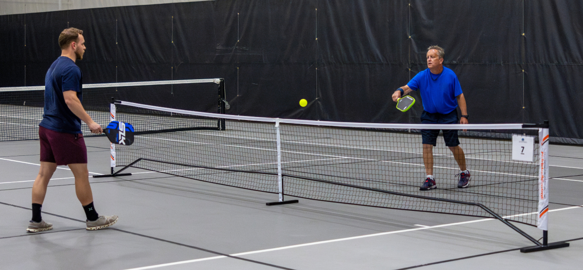 Two men playing pickleball on an indoor court, one hitting the ball over the net.