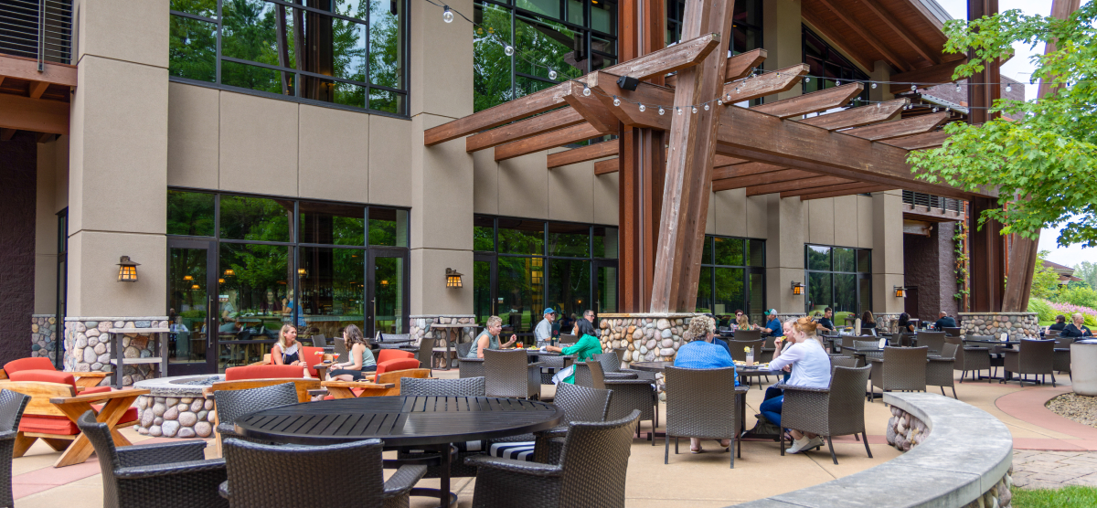 People enjoying an outdoor patio with tables, lounge seating, and a building in the background.