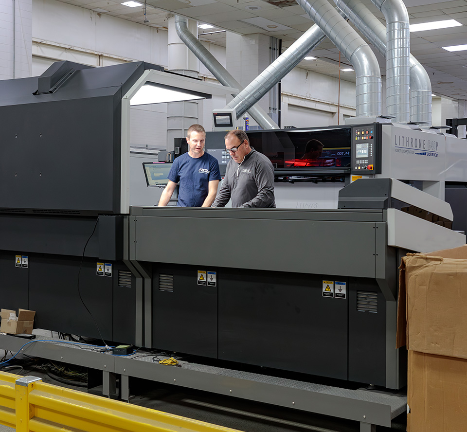 Two people working in printing factory with large scale printer.