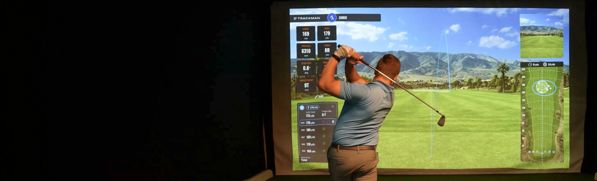 Man swinging a golf club in the SentryWorld golf simulator, with course projected on screen.