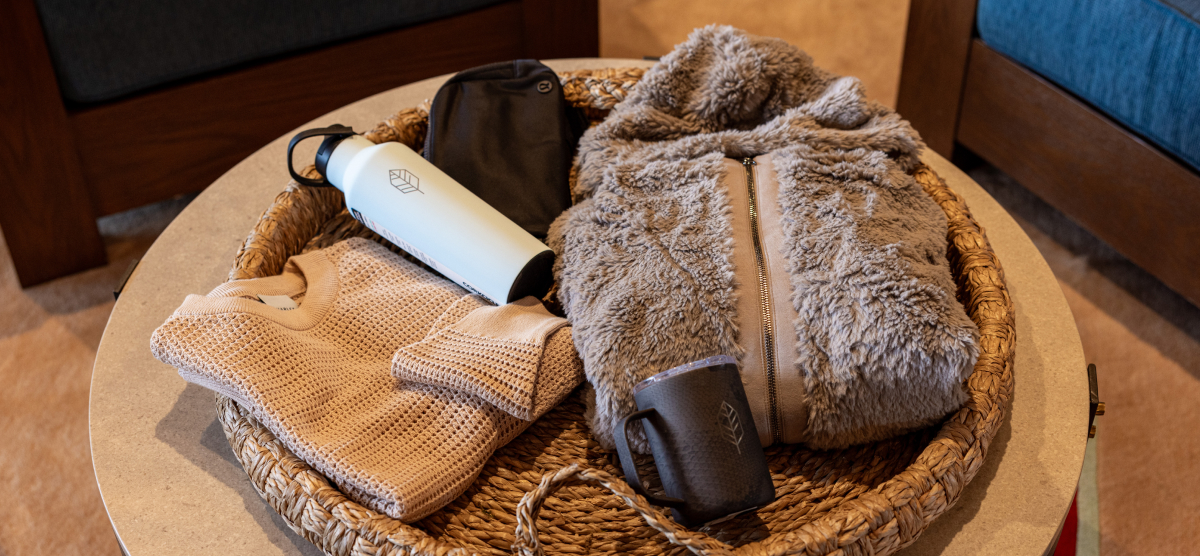 A woven basket on a table holding a water bottle, fuzzy jacket, sweater, and mug.