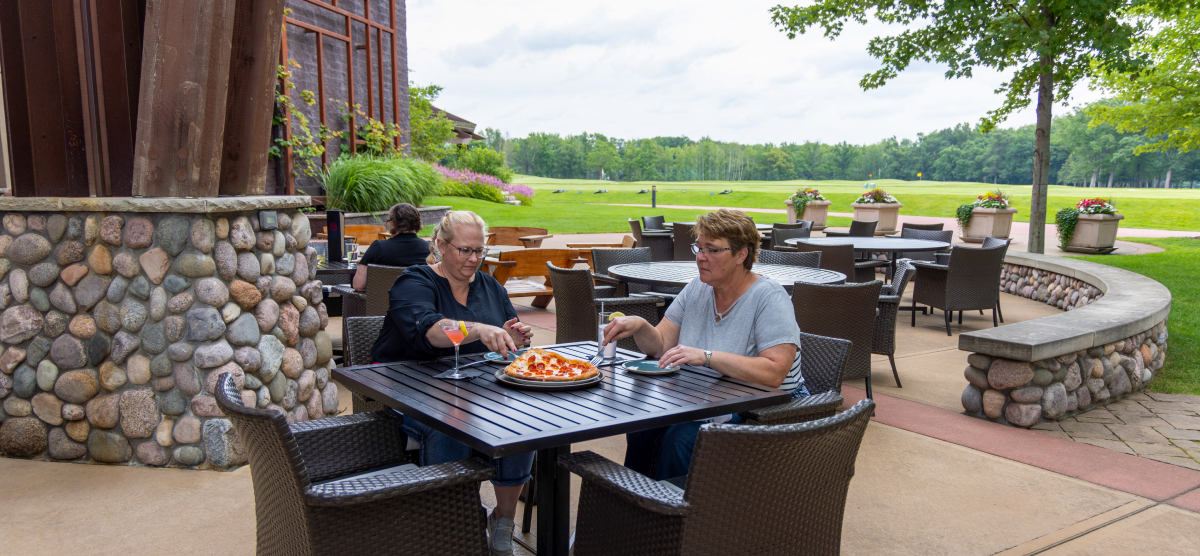 Two women eating pizza and drinking on an outdoor patio overlooking a golf course.