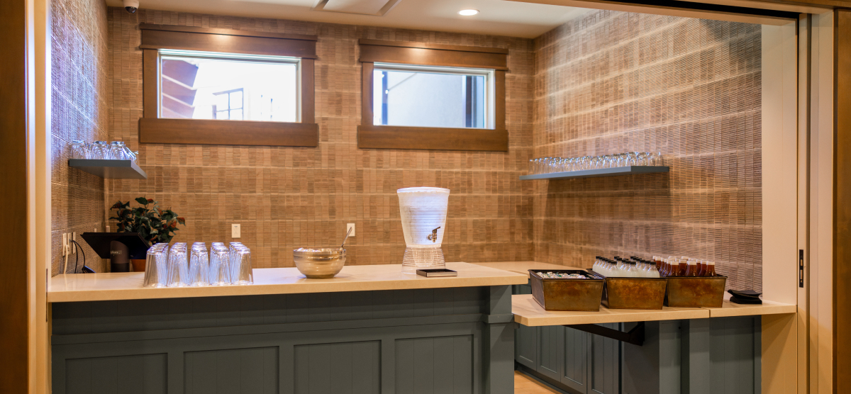 A bar area with a water dispenser, glasses, and drinks, against a textured wall with windows.