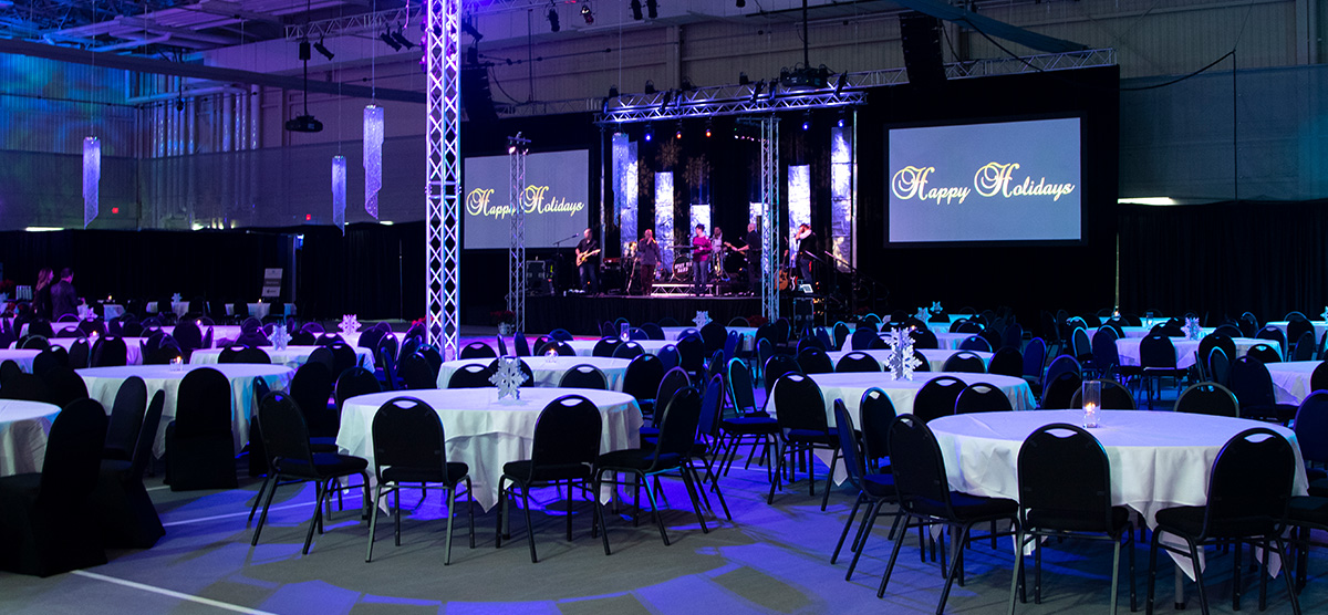 Tables and a stage with colorful lighting and a projector screen set for an event in the fieldhouse at SentryWorld