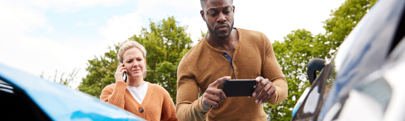 Man taking photo of car damage with phone, woman on phone looking concerned.