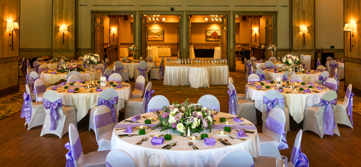 Elegant event room with round tables set with white linens, purple accents, and floral centerpieces.