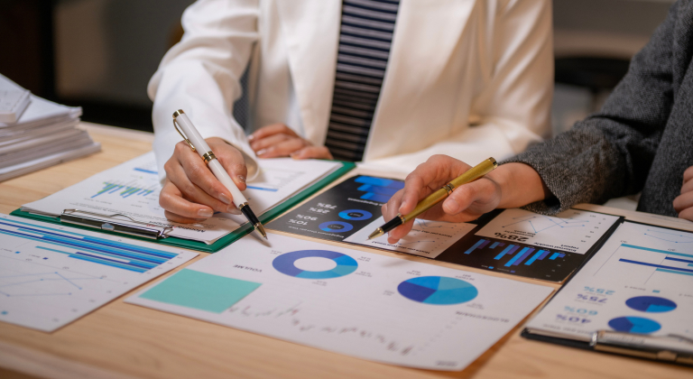 Two people’s hands with pens reviewing business documents with charts and graphs.