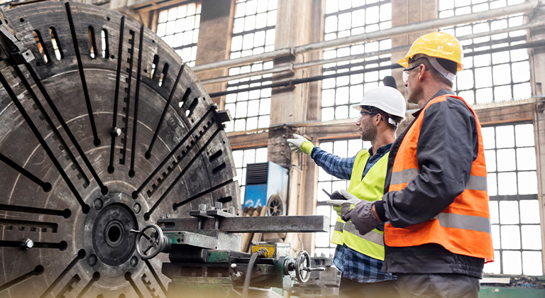 Two men in safety vests reviewing equipment on a manufacturing floor.