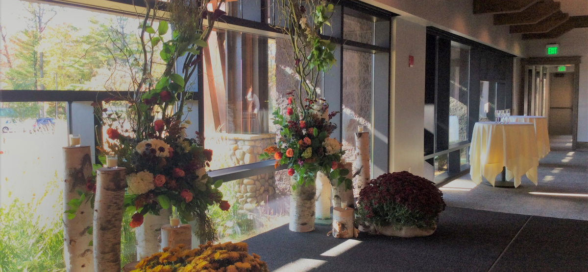 Indoor event space with large windows, floral arrangements, birch logs, and cocktail tables.