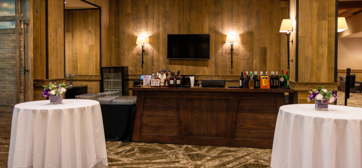 A wooden bar with bottles and a TV, flanked by two round tables with floral centerpieces.