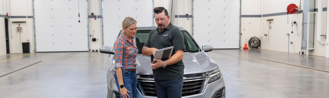 Two people looking at a laptop in front of a silver car in a garage.