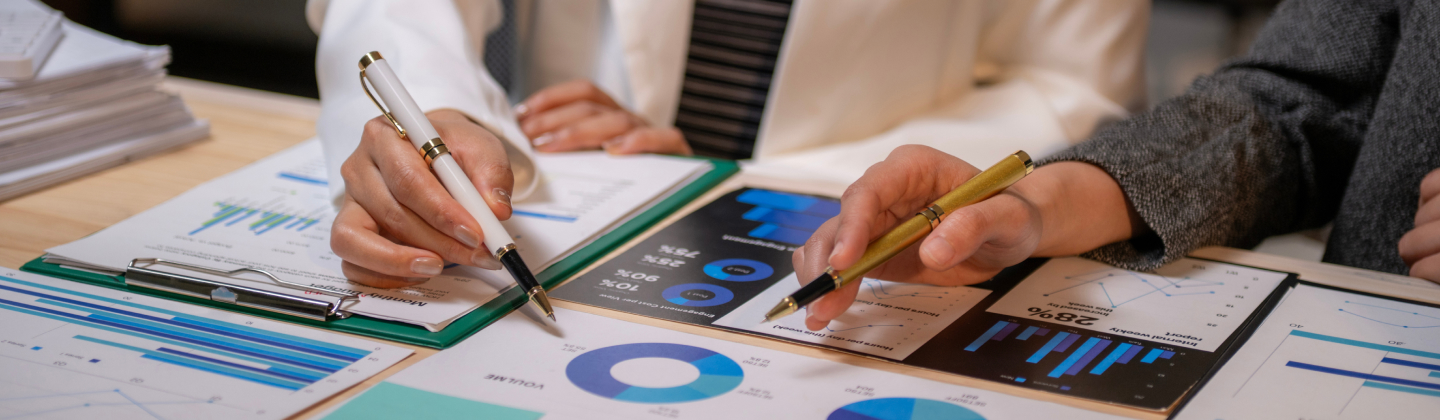 Two people’s hands with pens reviewing business documents with charts and graphs.