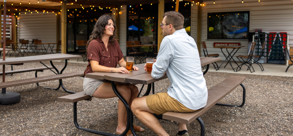 Two people sitting at an outdoor picnic table, enjoying drinks on a patio with string lights.