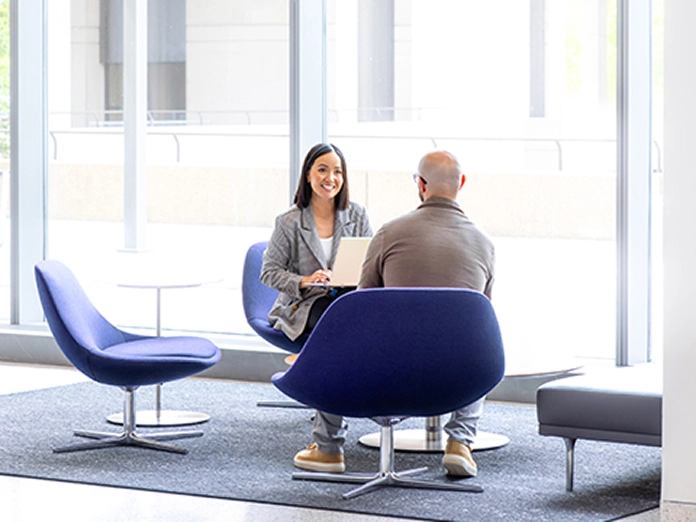 teams-claims-man-woman-sitting-talking-696x522 A woman with a computer talking with a man in a lobby