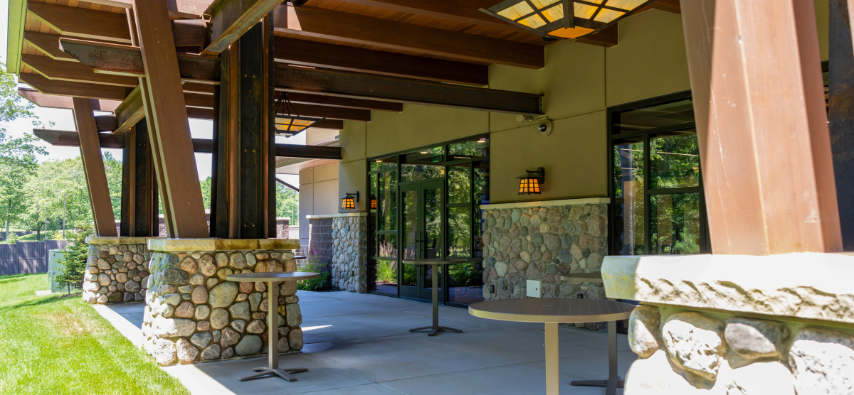 Outdoor patio with stone pillars, wooden beams, small tables, and glass doors to a building.