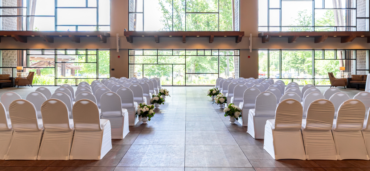 Event room set for a ceremony with rows of white-covered chairs and floral aisle decorations.