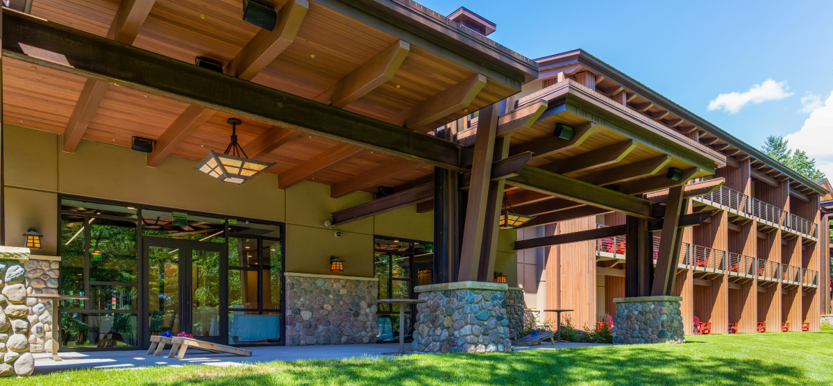 Building exterior with covered patio, stone pillars, wooden beams, and a cornhole game on the grass.