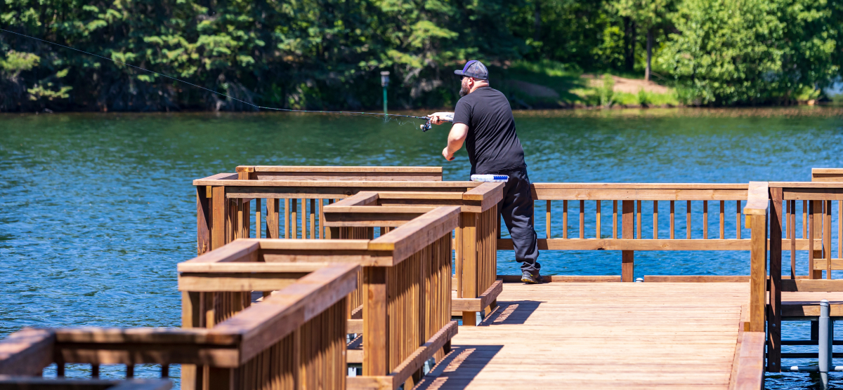 A man fishing from a wooden pier on a calm lake surrounded by green trees.