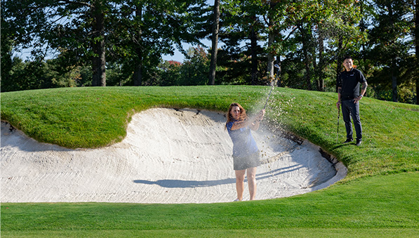 Golfer hitting her ball out of a sand trap while her golf partner watches. 
