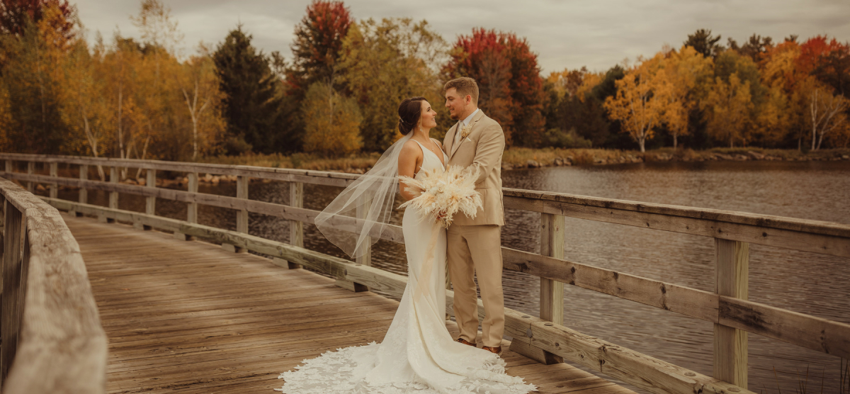 Bride and groom on a wooden bridge over water, with colorful autumn trees in background.