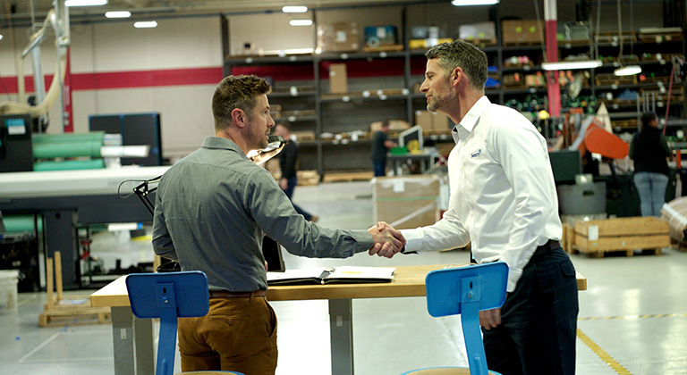 Two men shaking hands across a table in a busy warehouse or industrial facility.