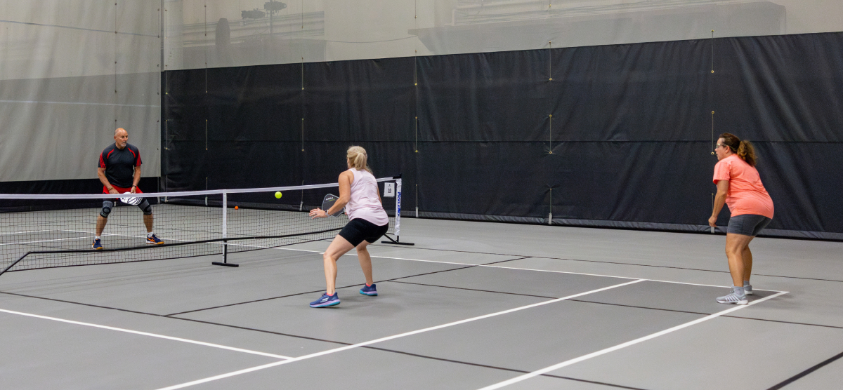 Three people playing pickleball on an indoor court, one man at the net, two women opposite.