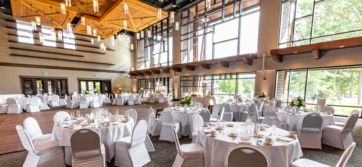 Large event room with many round tables set for a reception, featuring white linens and floral centerpieces.