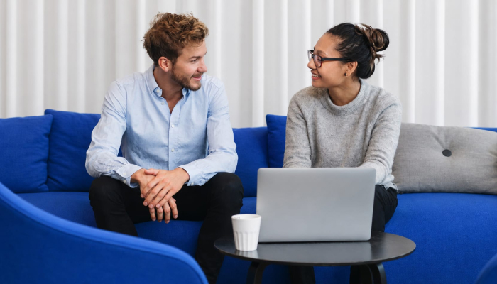 Two colleagues smiling and talking on a blue couch with a laptop.