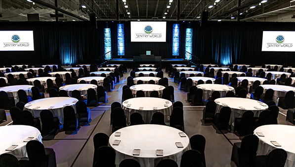 Meeting set up in the fieldhouse with round tables, stage and three monitors. 