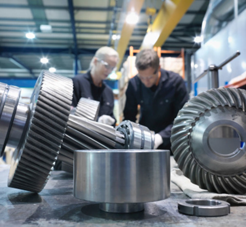 Metal parts in foreground with two metalworkers behind.