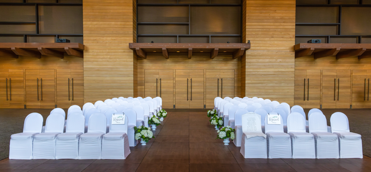 Indoor ceremony setup with rows of white chairs, an aisle, and floral decorations.