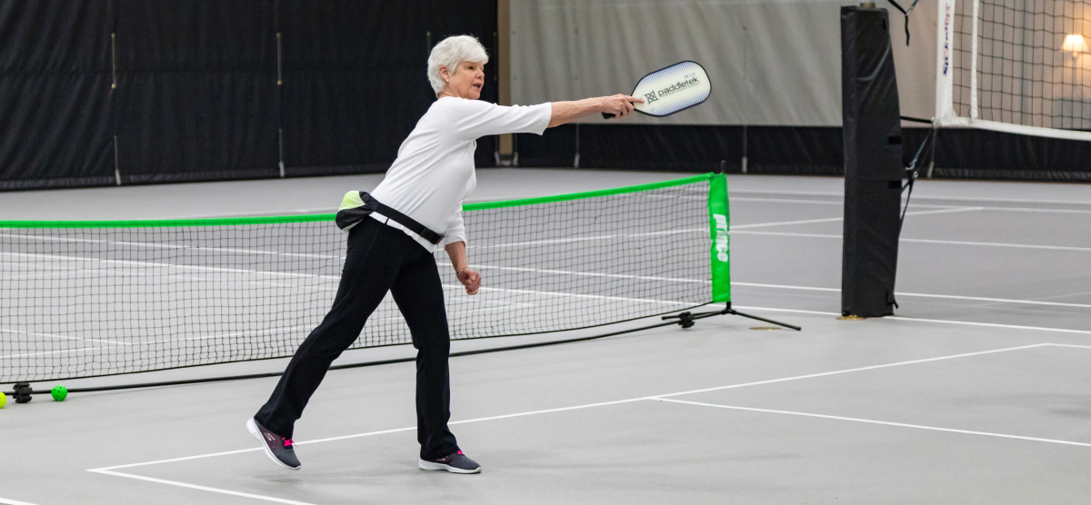 Woman in white shirt and black pants playing pickleball on an indoor court, paddle extended.