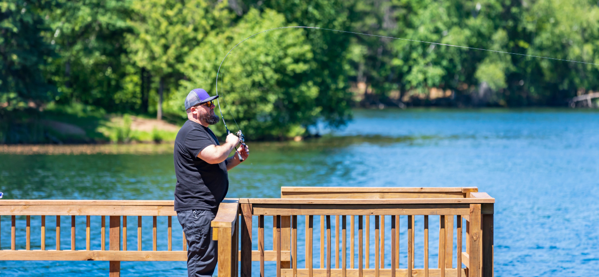 A man in a hat and t-shirt fishing from a wooden dock on a lake.