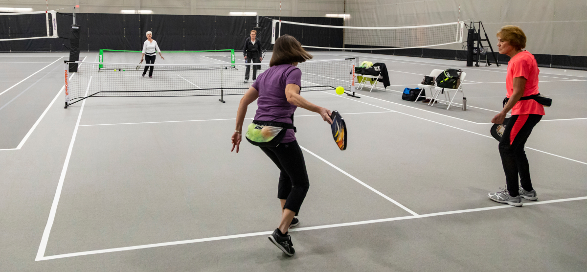 Four women playing pickleball on an indoor court, two in the foreground with paddles.