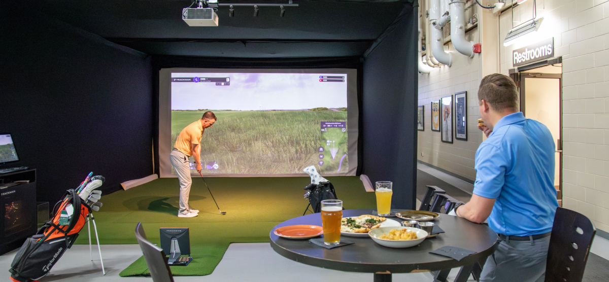 Two men in a golf simulator, one playing golf, the other eating pizza and drinking beer.